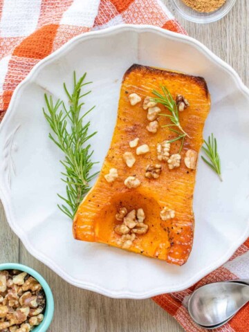 White plate with a baked half of butternut squash covered in nuts and rosemary sprigs.