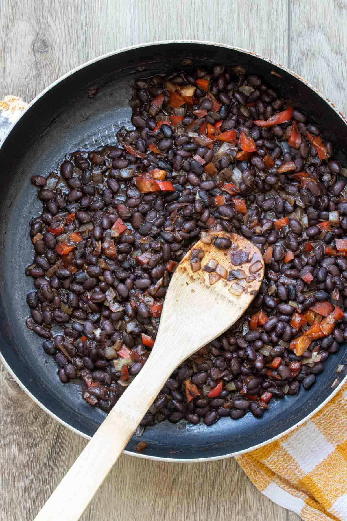 Wooden spoon mixing black beans, red peppers and onions in a pan.