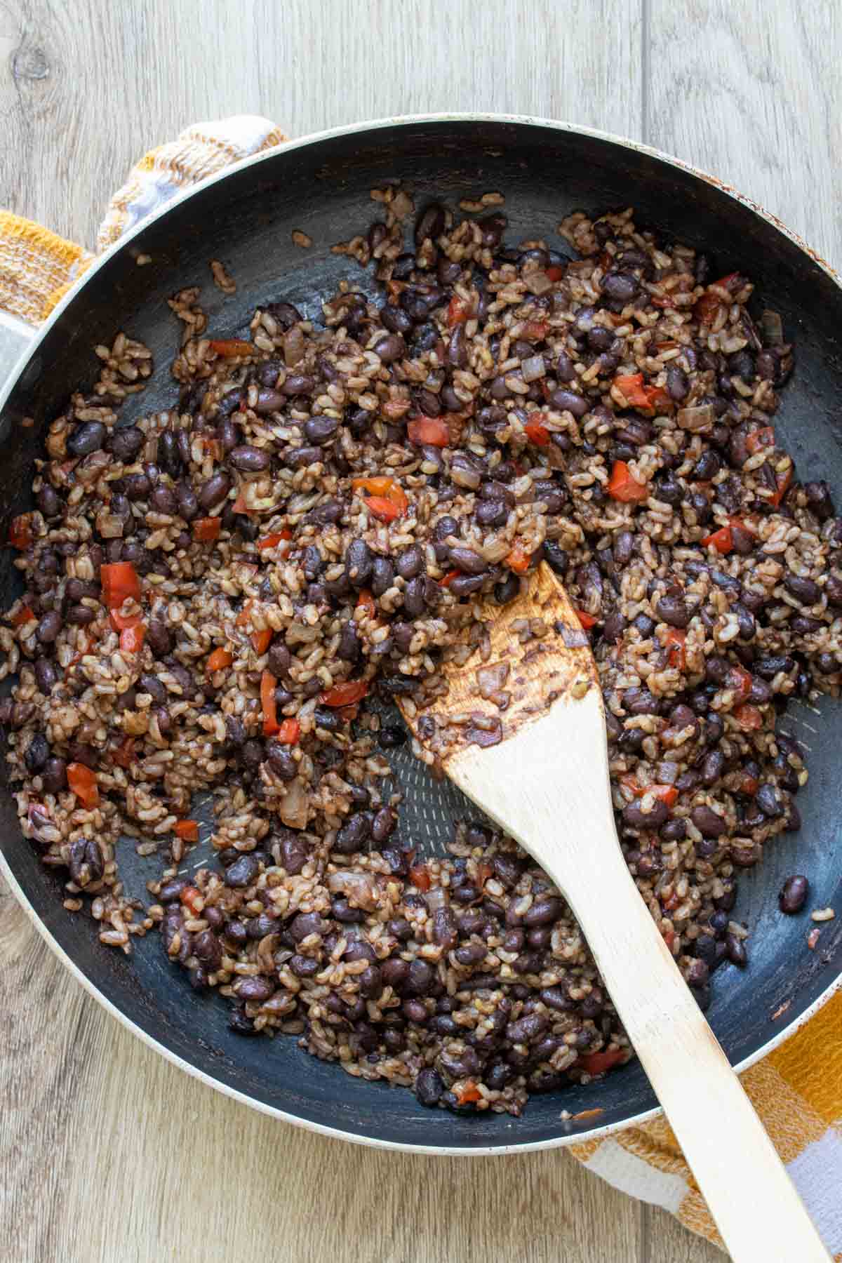 Rice, black beans, and chopped red pepper being stirred in a pan by a wooden spoon.