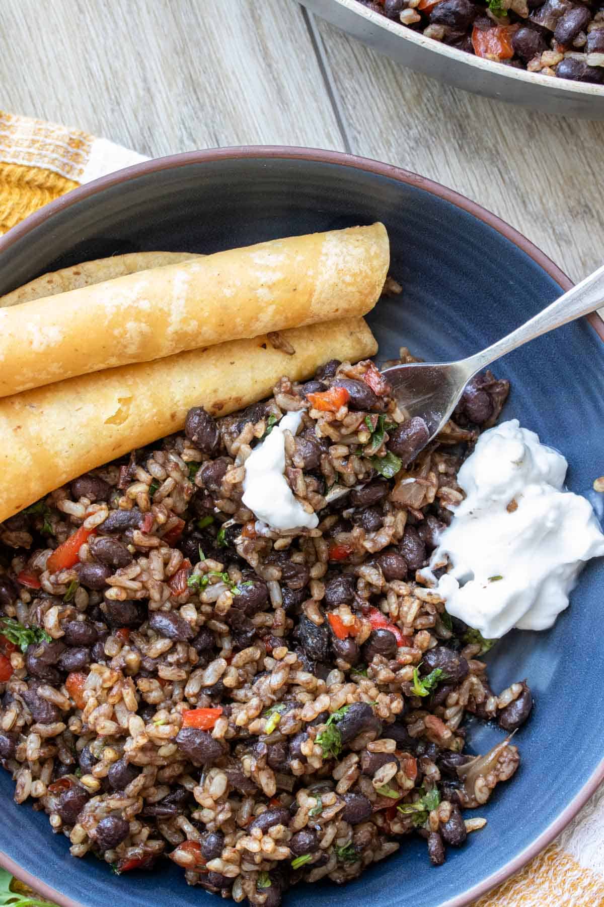 Fork getting a bite of a black bean and rice mix from a blue bowl with corn tortillas and sour cream.