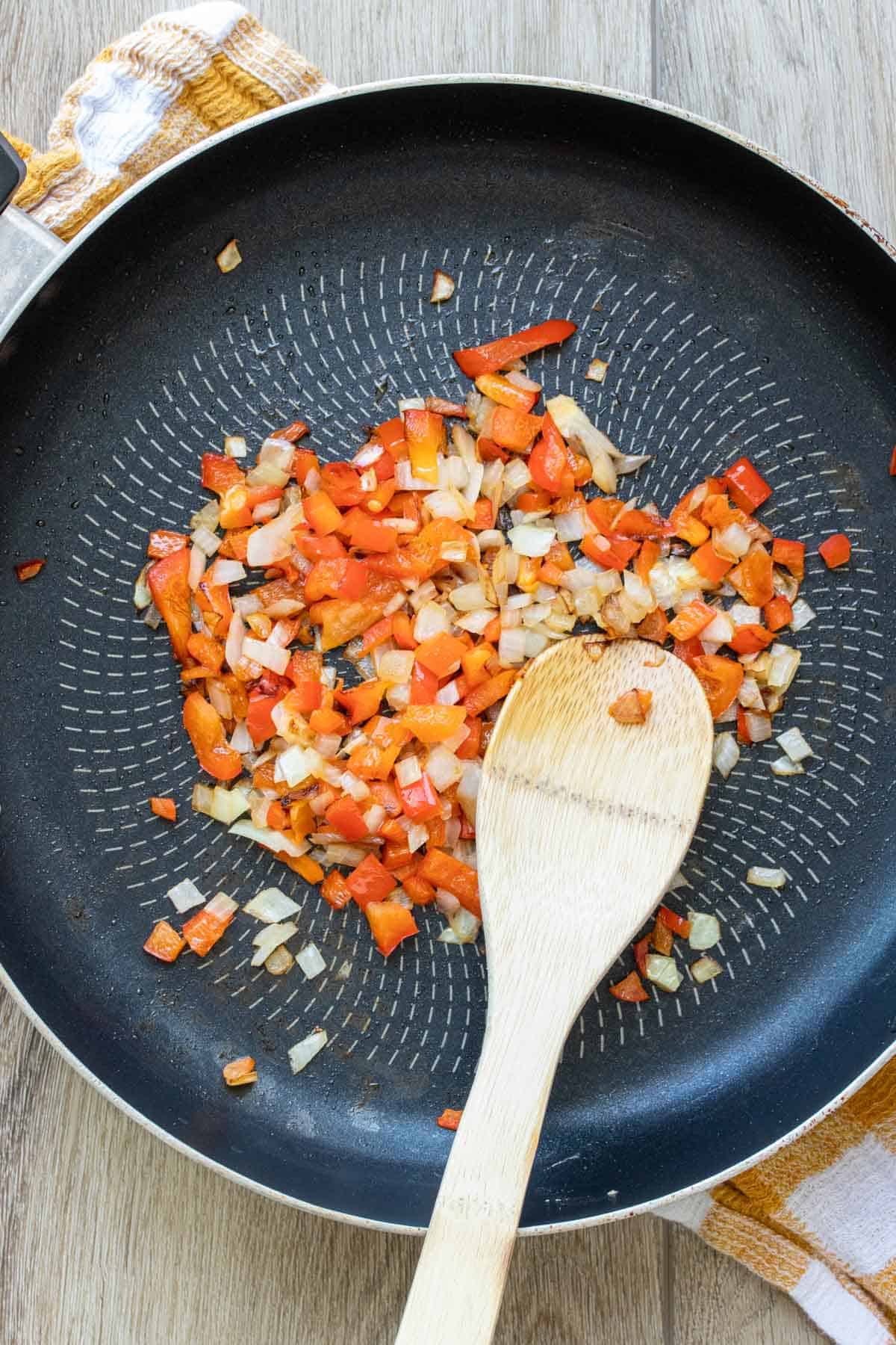 Red peppers and onions being sauteed in a pan with a wooden spoon