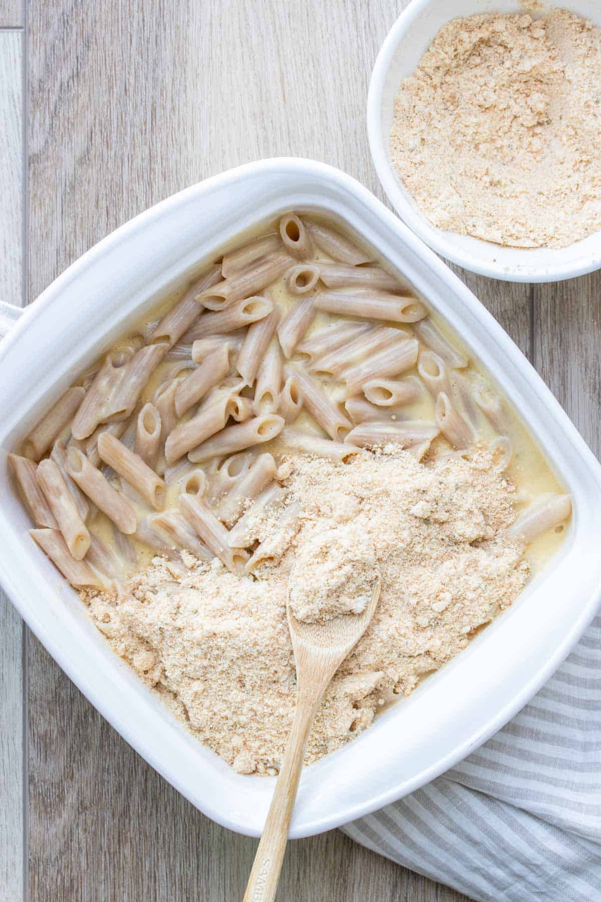 Pasta mixed with a creamy sauce in a white baking dish being covered by crispy topping