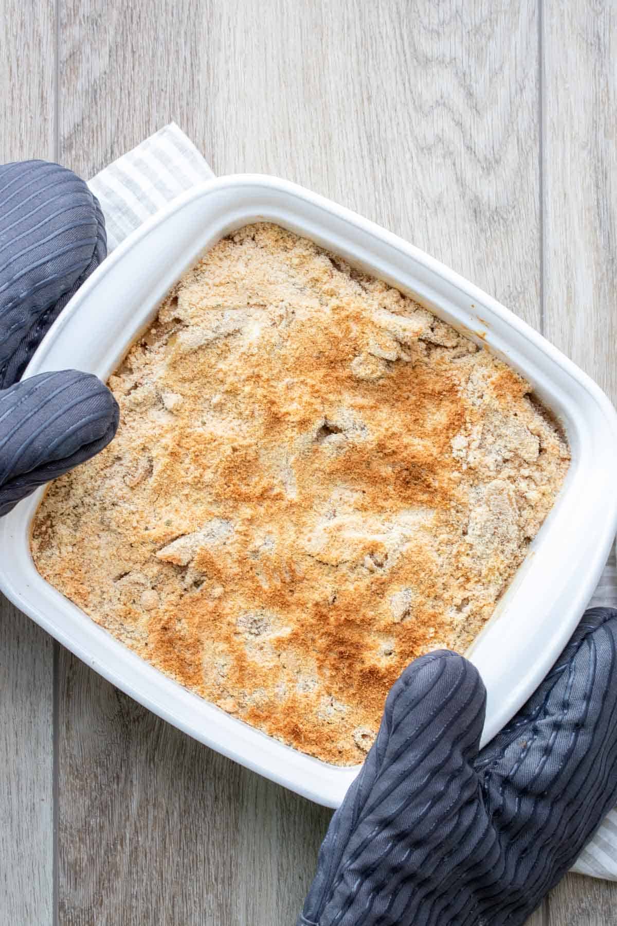 Potholders holding a white baking dish with a baked pasta and crispy topping in it