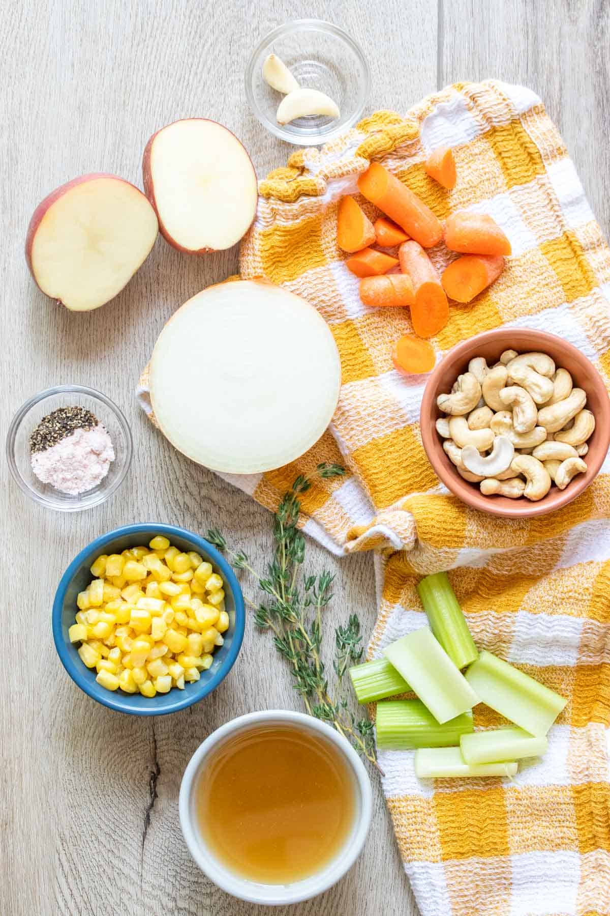 Ingredients needed to make corn chowder with a cashew base in bowls and on a wooden surface with a checkered towel