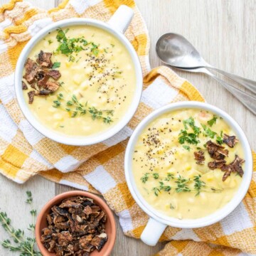 Two soup bowls on a yellow and white checkered towel filled with corn chowder next to toppings on a wooden surface