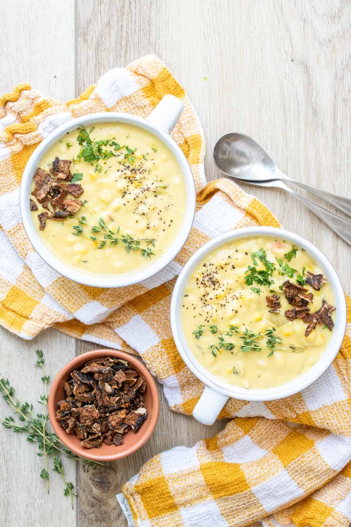 Two soup bowls on a yellow and white checkered towel filled with corn chowder next to toppings on a wooden surface