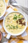 White soup bowl with corn chowder topped with herbs and bacon bits sitting on a checkered towel next to spoons