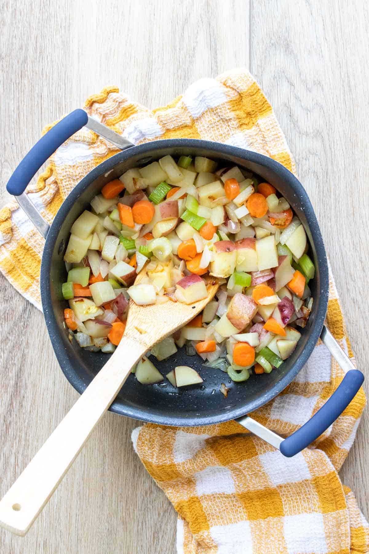 Celery, carrots, potatoes and corn in a pot being mixed by a wooden spoon