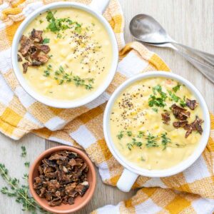 Two white soup bowls with corn chowder and topped with herbs and bacon bits