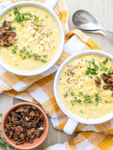 Two white soup bowls with corn chowder and topped with herbs and bacon bits