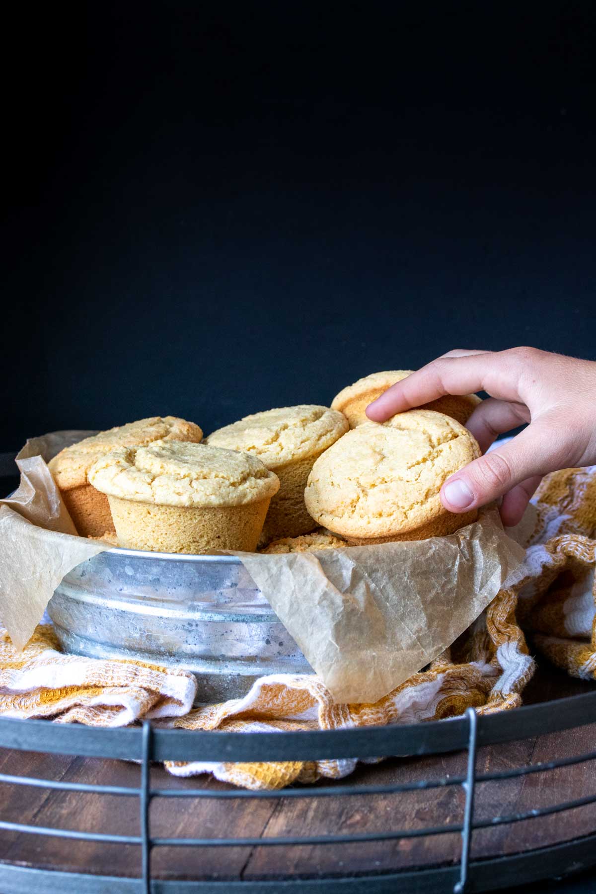 Hand grabbing a yellow muffin from a metal bowl on a yellow checkered towel