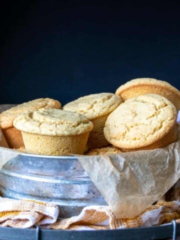 Metal tin on a checkered towel piled up with yellow cornbread muffins in parchment paper