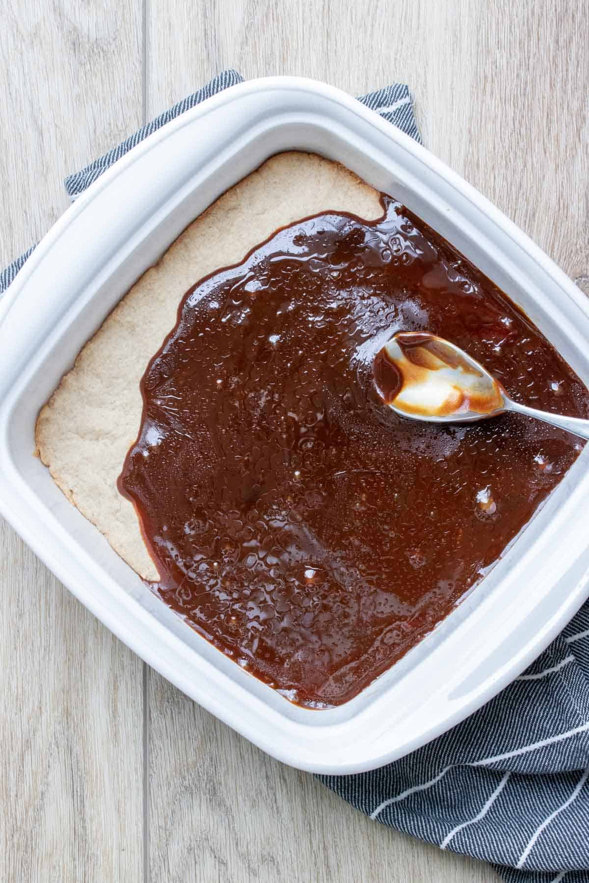 A spoon spreading caramel over baked shortbread in a square white baking dish