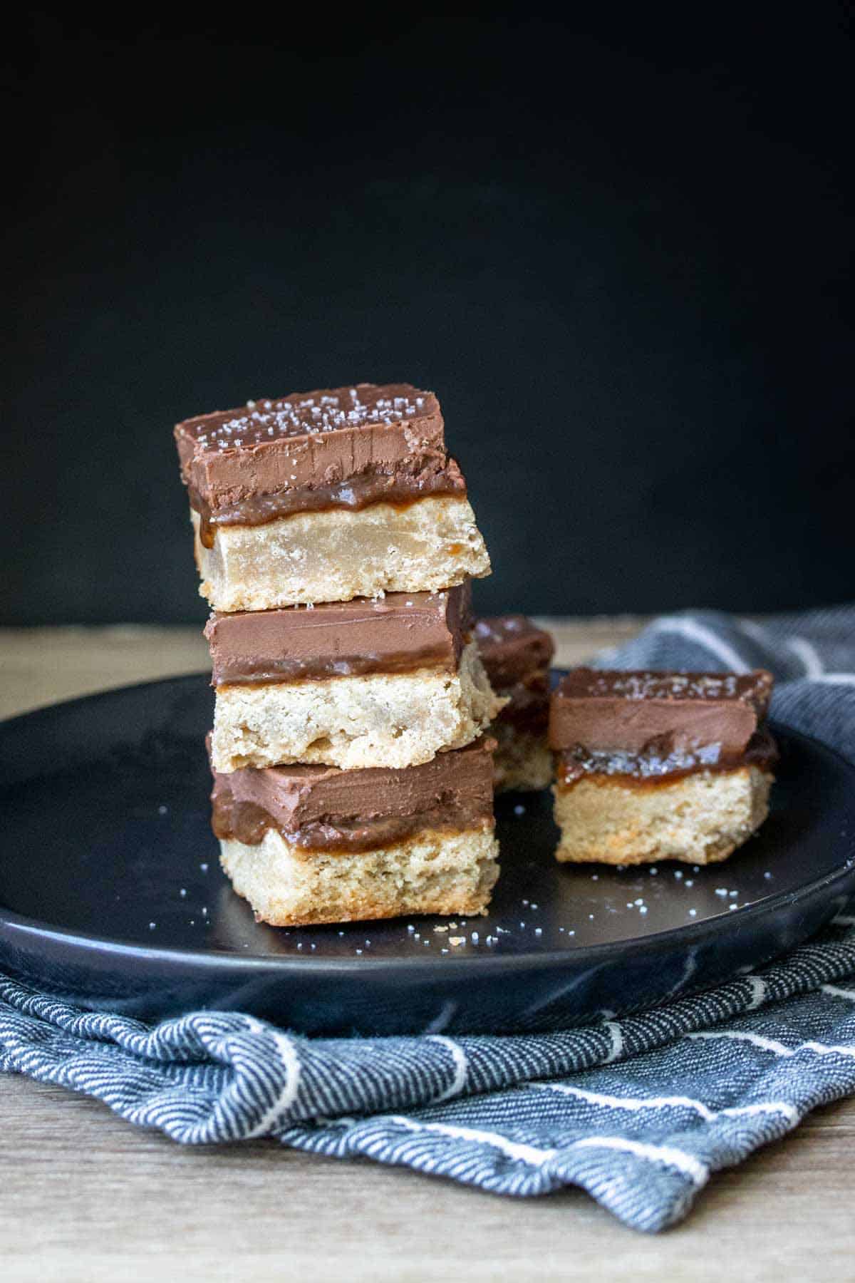 A black plate stacked with squares of millionaire shortbread dessert sprinkled with sea salt