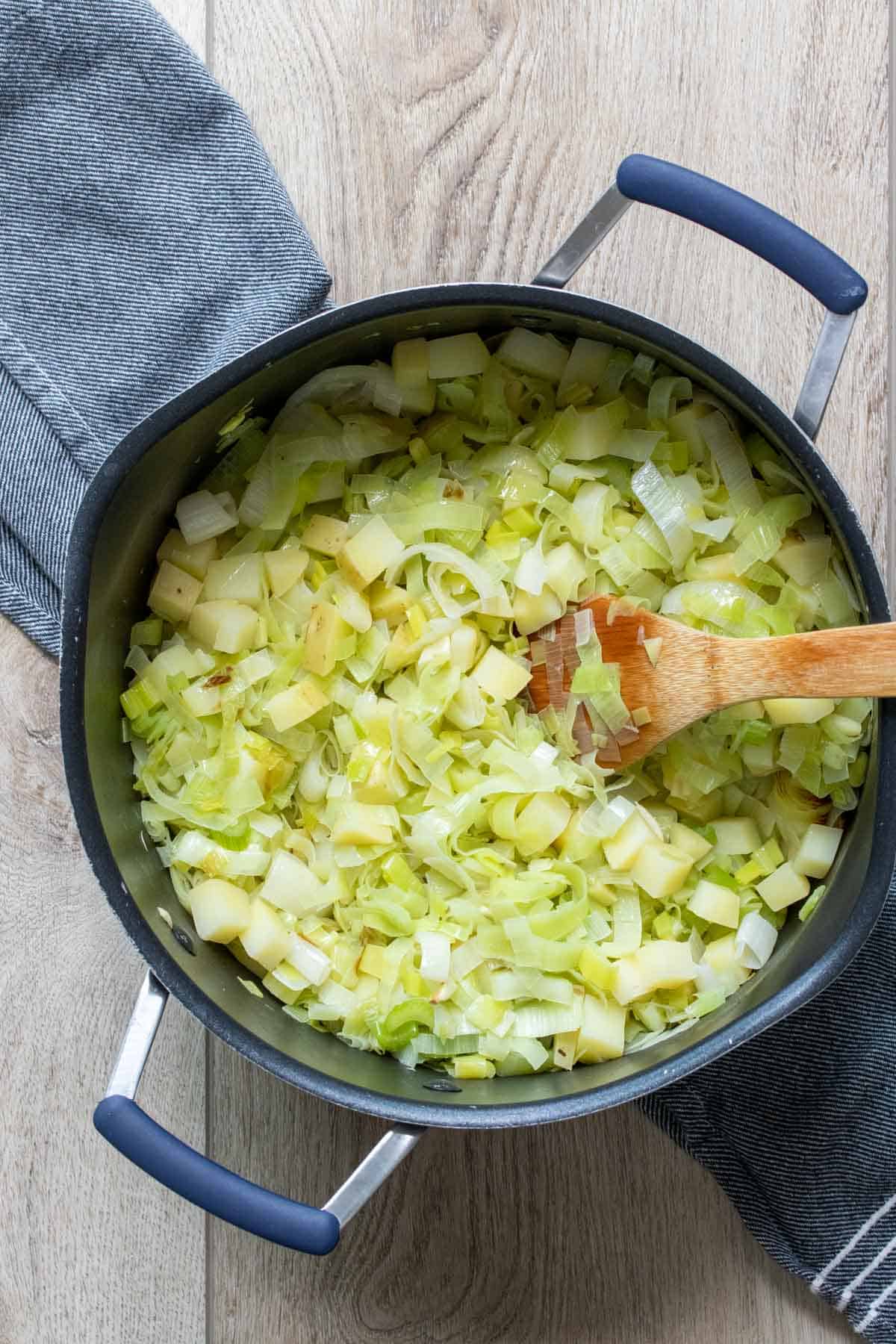 Wooden spoon mixing chopped leeks and potatoes in a black pot on a wooden surface