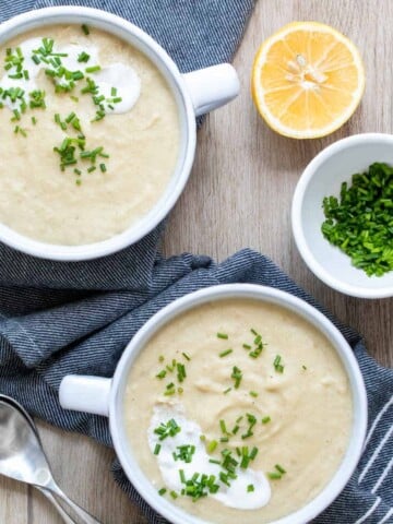 Two white soup bowls with pureed soup next to a cut lemon and white bowl of chives