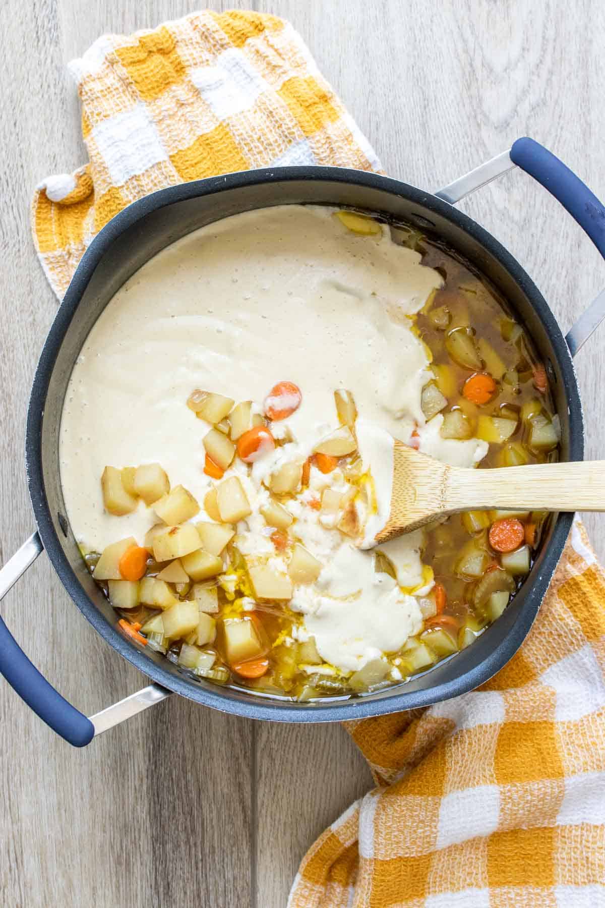 Wooden spoon mixing cream into a bowl of broth with veggies
