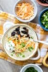 A cream bowl with loaded potato soup and a spoon in it next to bowls of toppings on a yellow towel