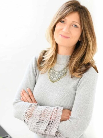 A woman with long blond hair and a grey sweater crossing her arms and smiling