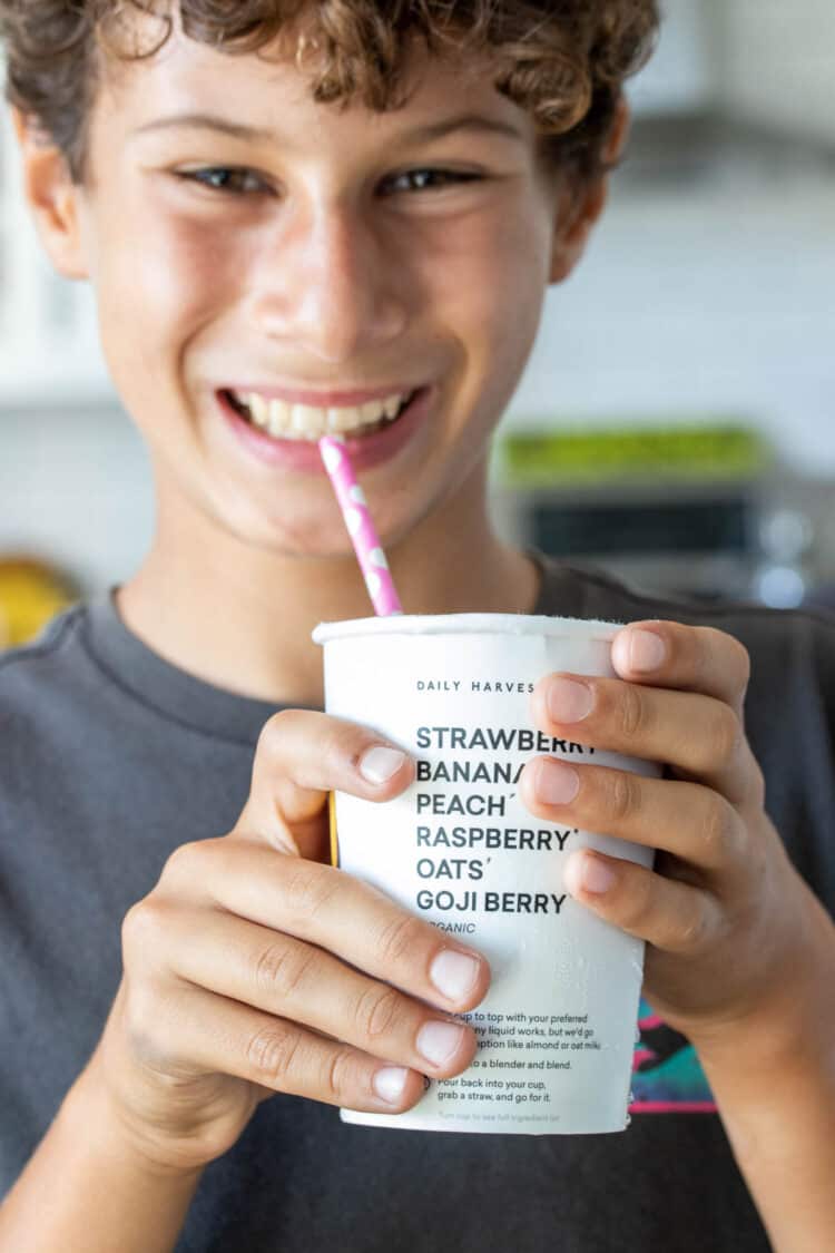 Photo of a boy smiling, holding a white paper cup with black writing and drinking through a straw