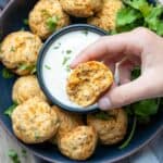 A half eaten sausage and cheese ball being held over a dark blue bowl with more balls and white dip.