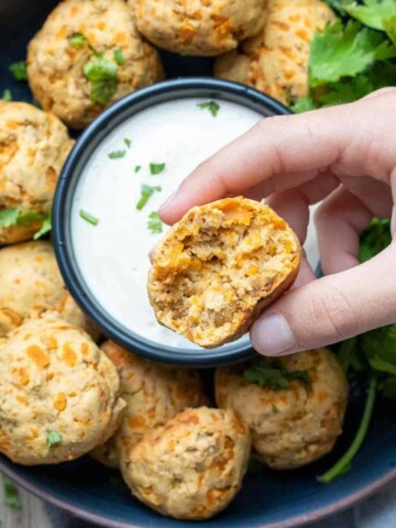 A half eaten sausage and cheese ball being held over a dark blue bowl with more balls and white dip.