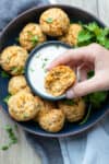 A photo of a hand holding a sausage ball that is bitten over a plate of them with a bowl of dip in the center.