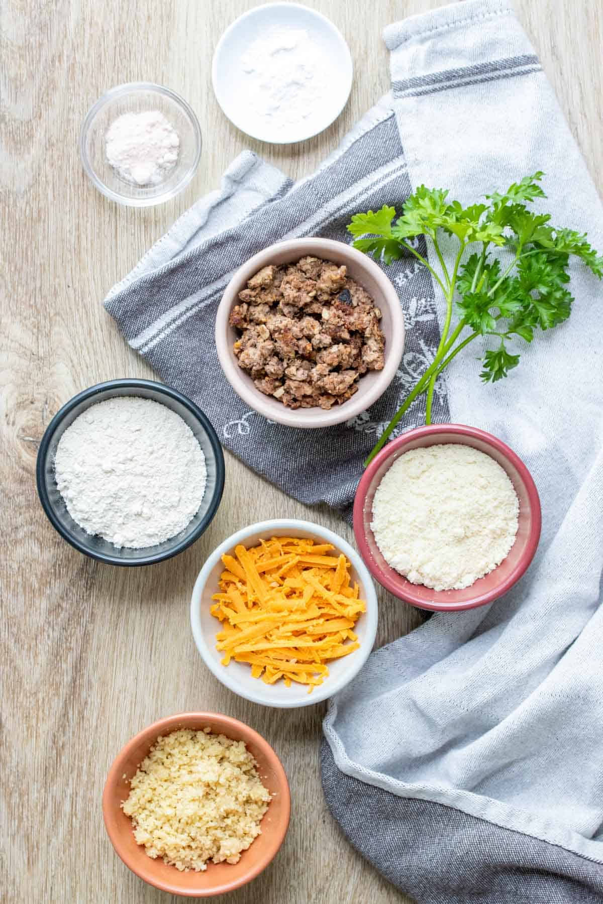 Bowls with flours, cheese, crumbled sausage baking powder and salt on a wooden surface with parsley.