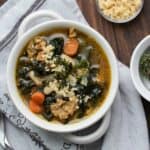 White bowl with a kale, sausage, carrot and white bean soup inside on a grey towel.