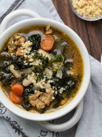 White bowl with a kale, sausage, carrot and white bean soup inside on a grey towel.