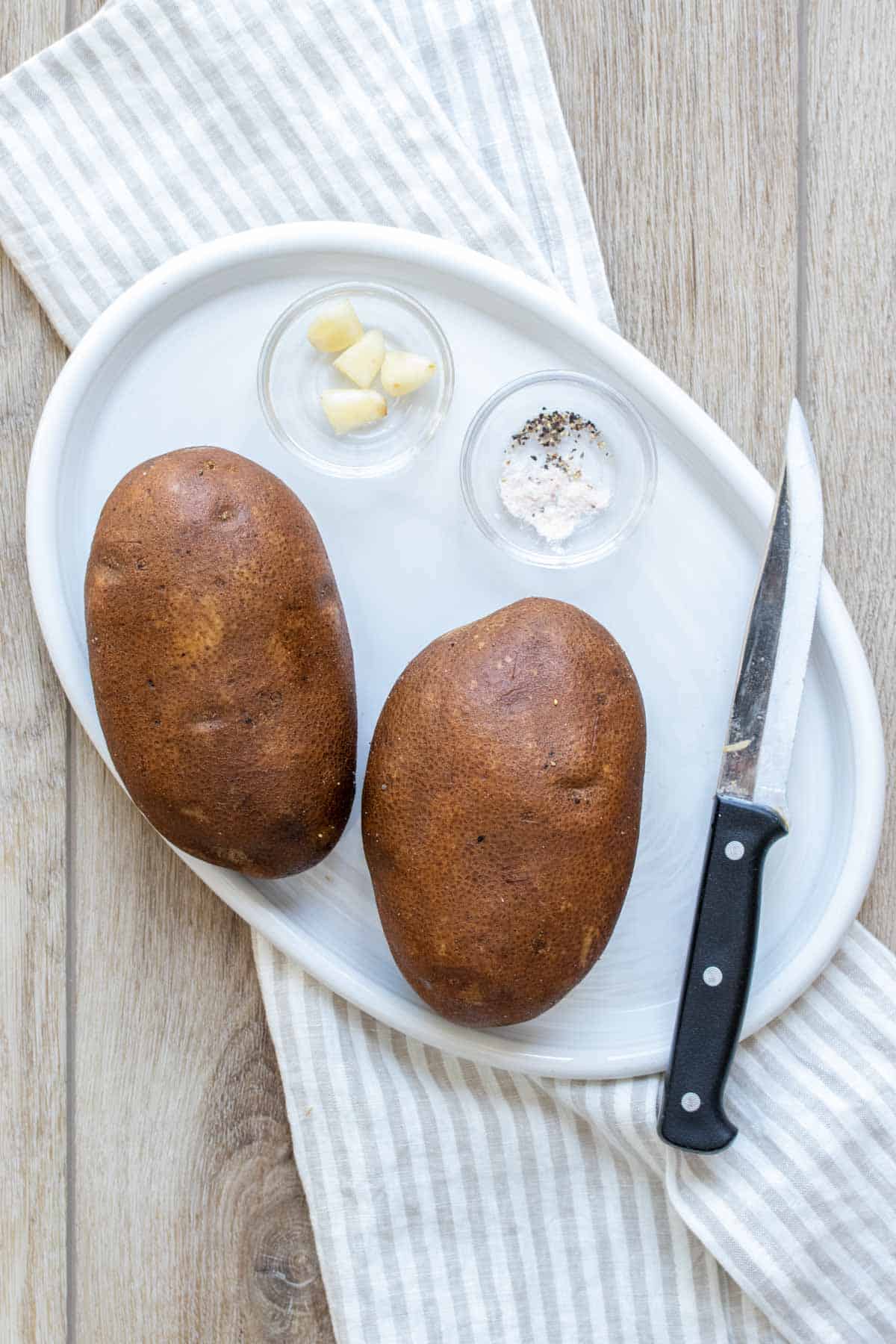 White plate with two whole potatoes, garlic, salt and pepper and a knife