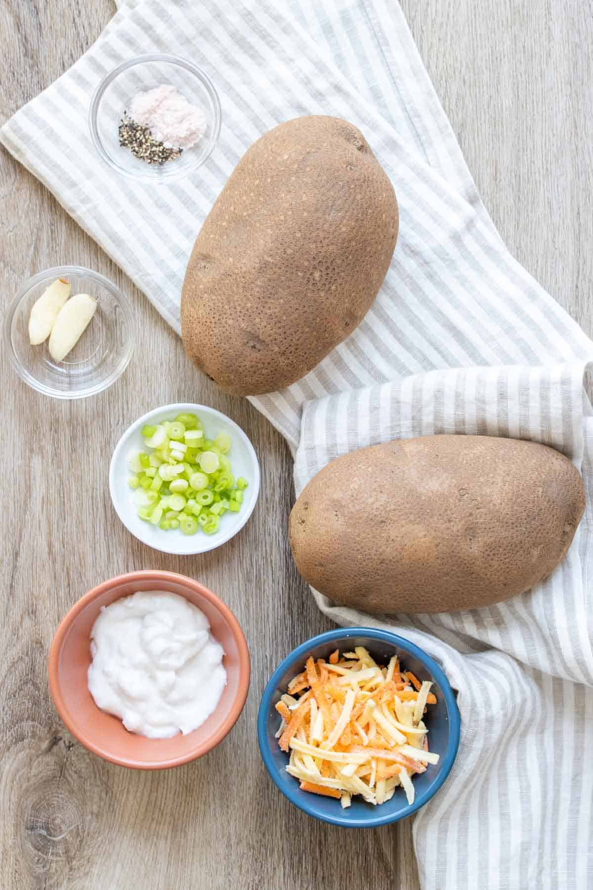 Whole potatoes, with bowls of cheese, sour cream, green onions, garlic, salt and pepper on a wooden surface