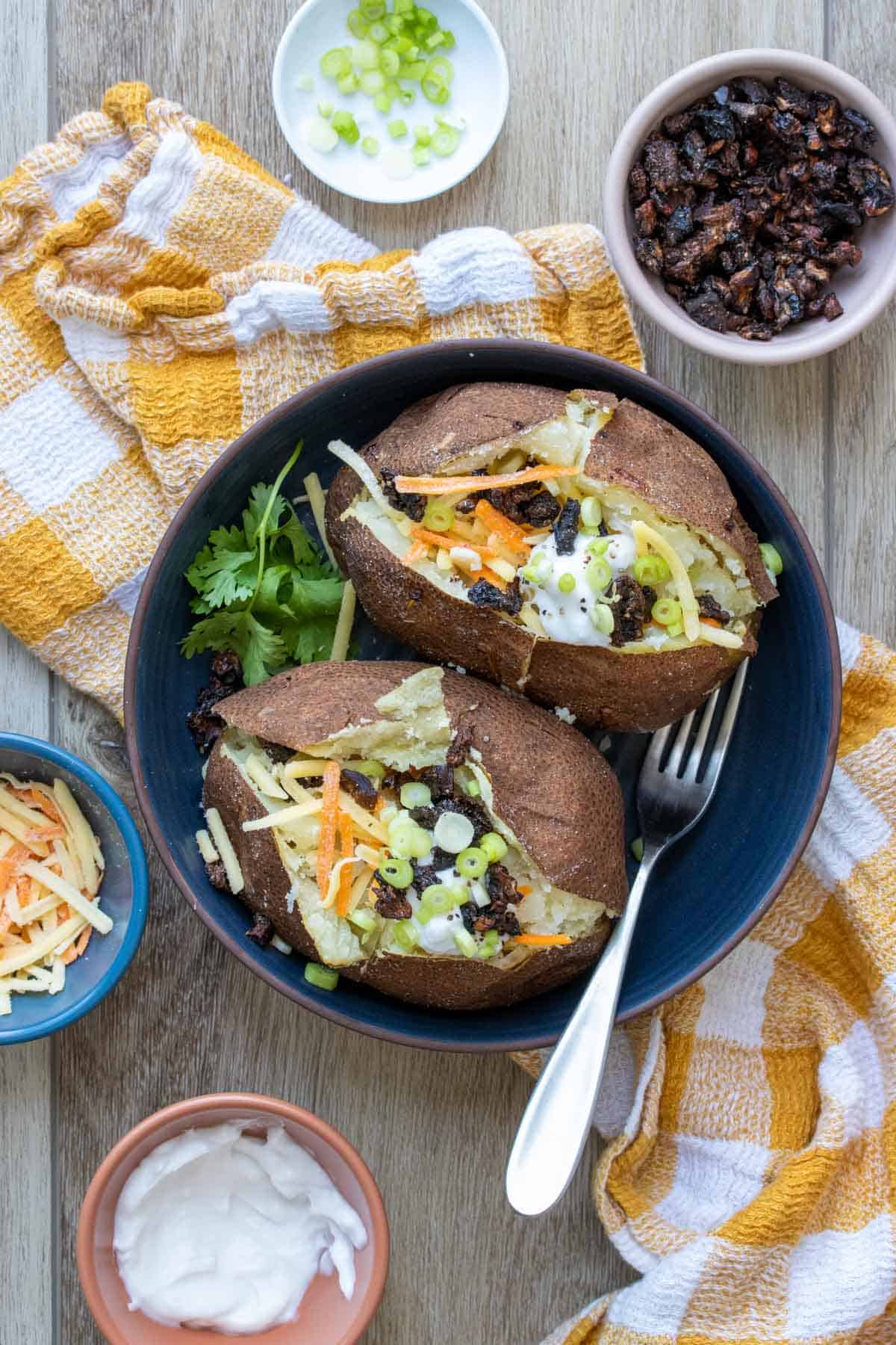 Blue bowl with two baked potatoes covered with toppings on a yellow checkered towel