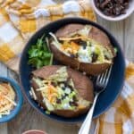 Blue bowl with loaded baked potatoes on a checkered towel surrounded by toppings