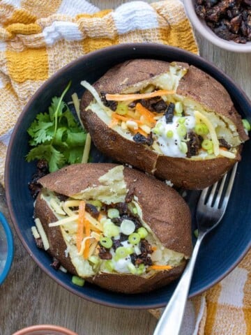 Blue bowl with loaded baked potatoes on a checkered towel surrounded by toppings