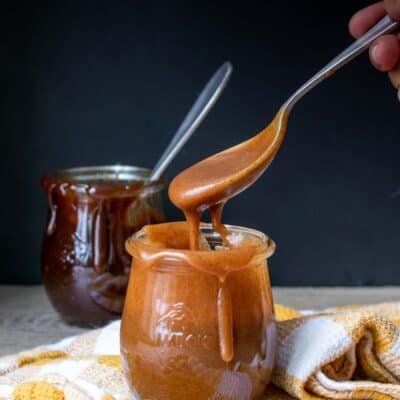 Hand holding a spoon with a light caramel sauce dripping off of it into a jar