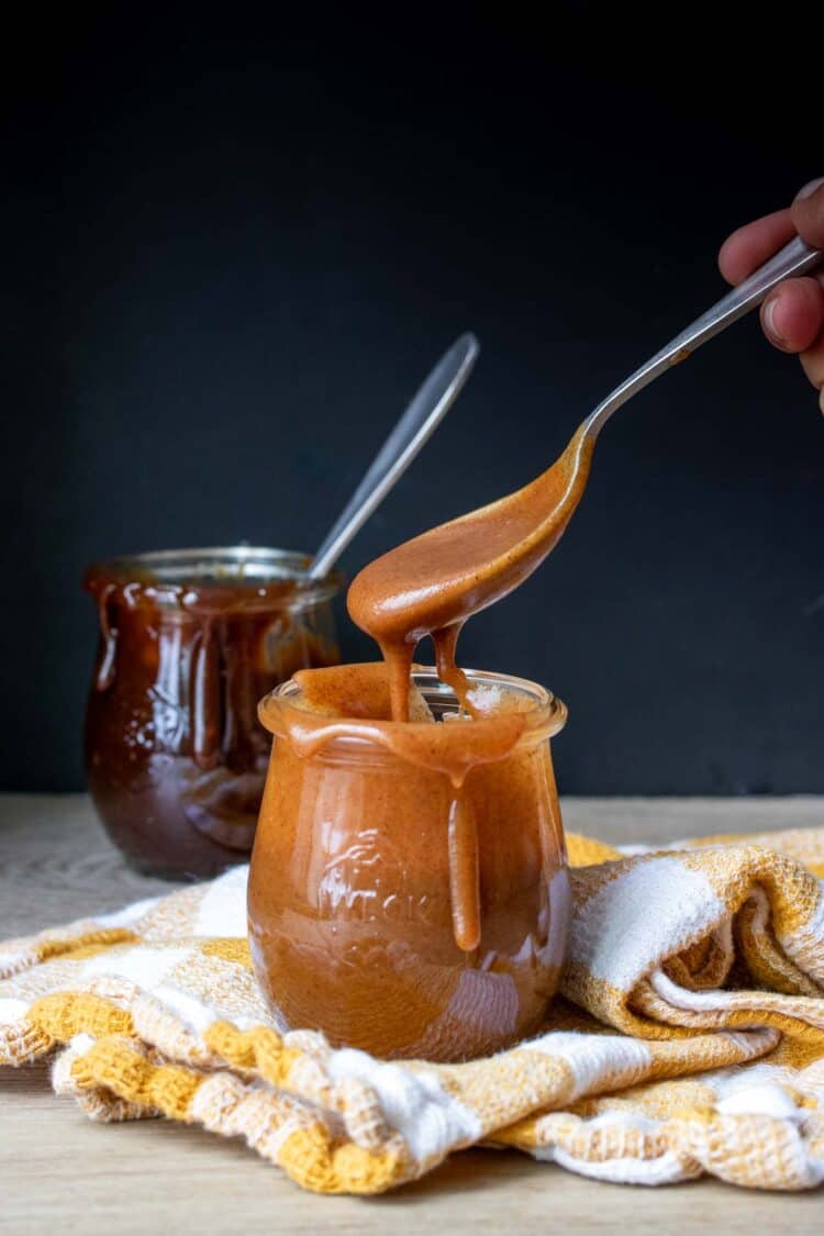Hand holding a spoon with a light caramel sauce dripping off of it into a jar
