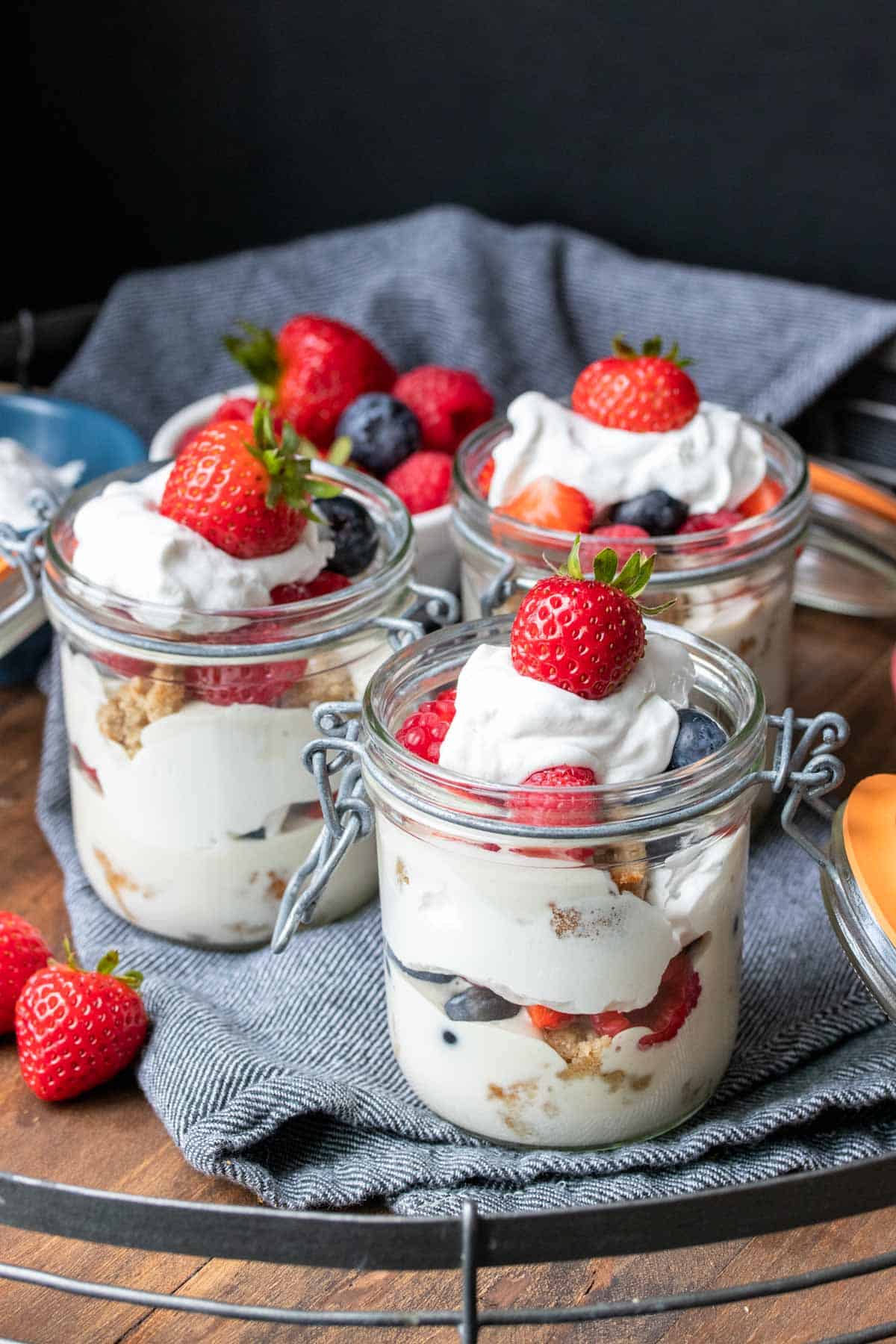 Three glass jars layered with cream, pudding, cake and berries on a wooden tray