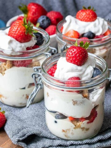 Three glass jars with hinged lids layered with cake, whipped cream, pudding and berries on a dark blue towel