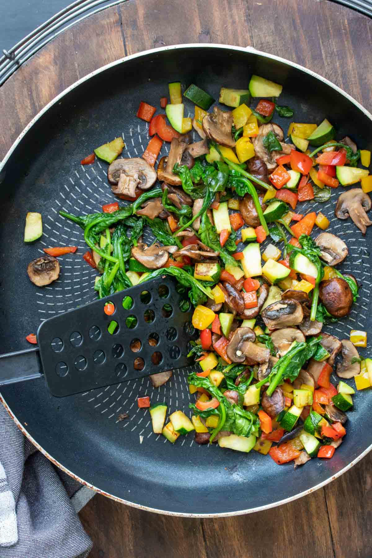 Spatula mixing chopped mushrooms, peppers, zucchini and spinach in a pan