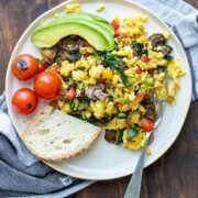 Grey plate with scrambled eggs mixed with veggies, slice of bread, avocado slices and cherry tomatoes