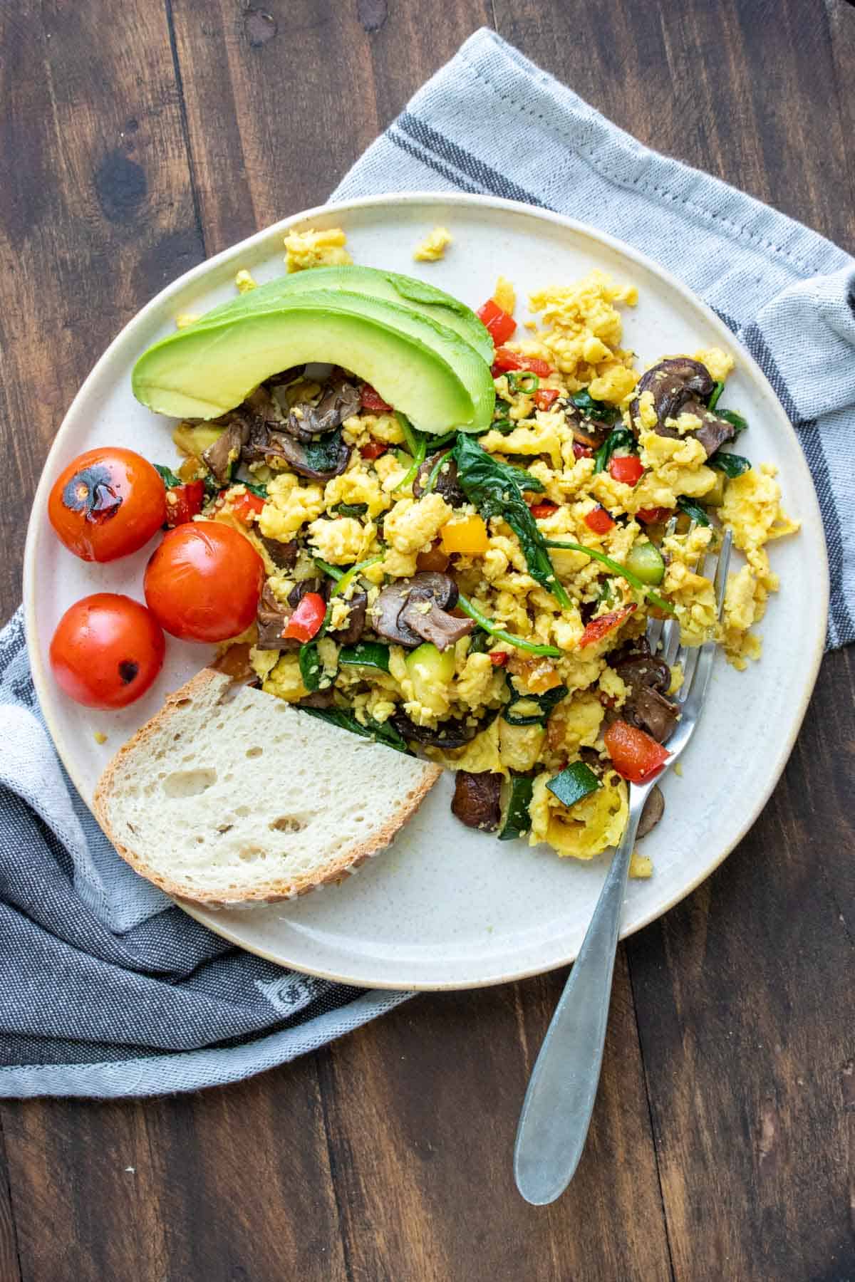 Grey plate with scrambled eggs mixed with veggies, slice of bread, avocado slices and cherry tomatoes