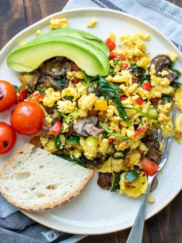 Veggie scrambled eggs on a plate with avocado slices, cherry tomatoes and a slice of bread on a grey towel
