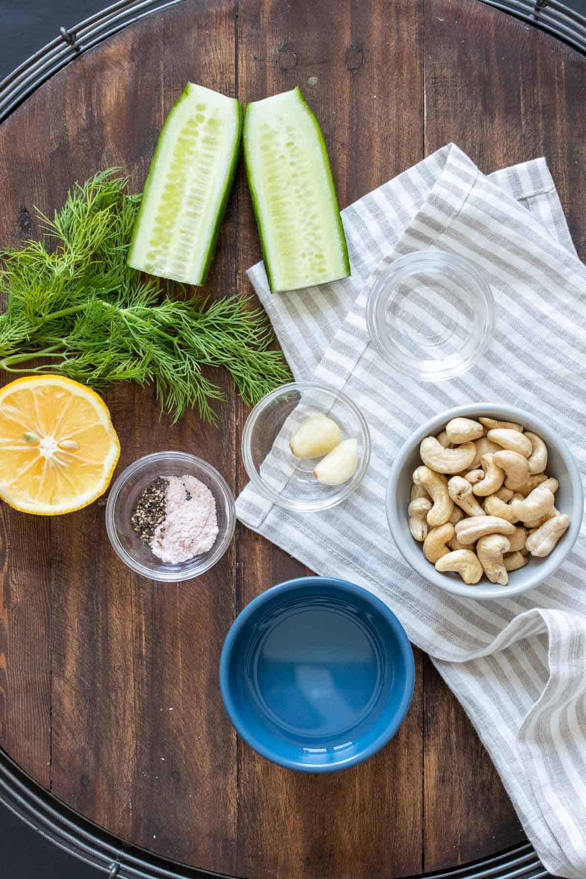 Bowls with cashews, water, salt, pepper, garlic and white vinegar on a wooden tray with cucumber, lemon and dill