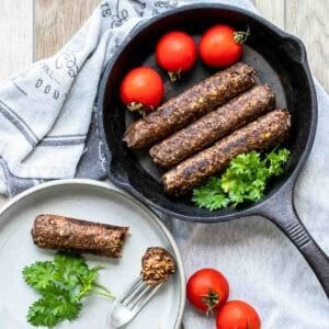 A sausage on a plate being eaten next to three sausages in a cast iron skillet.