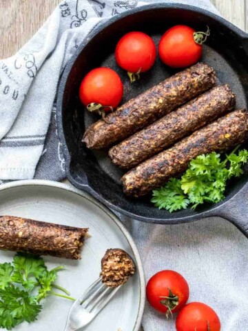 A sausage on a plate being eaten next to three sausages in a cast iron skillet.