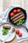 A cast iron pan with three sausages in it next to tomatoes and herbs and one cut on a plate next to it.