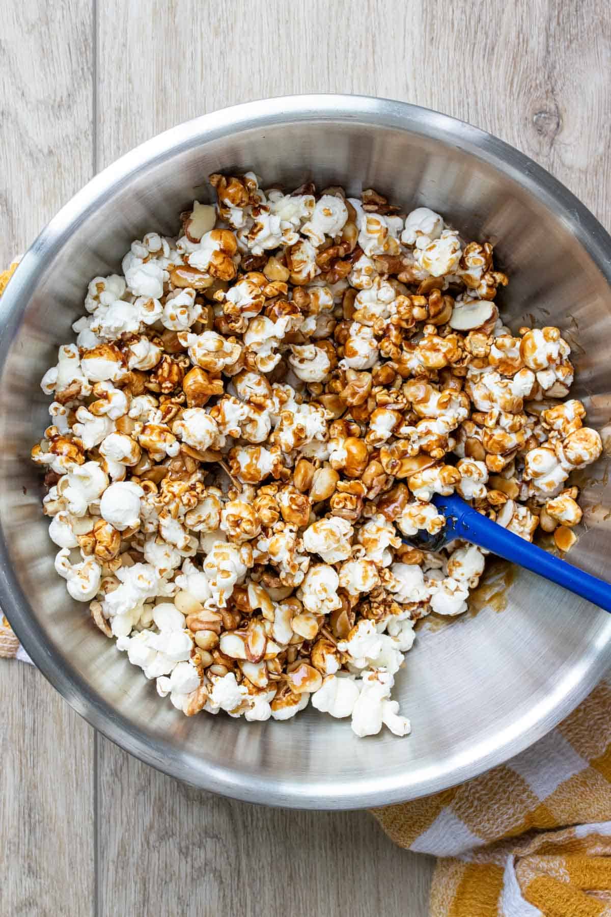 A metal bowl with popcorn being mixed with caramel by a blue spatula