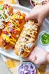 A hand holding a carrot dog in a bun covered in toppings near a plate with more