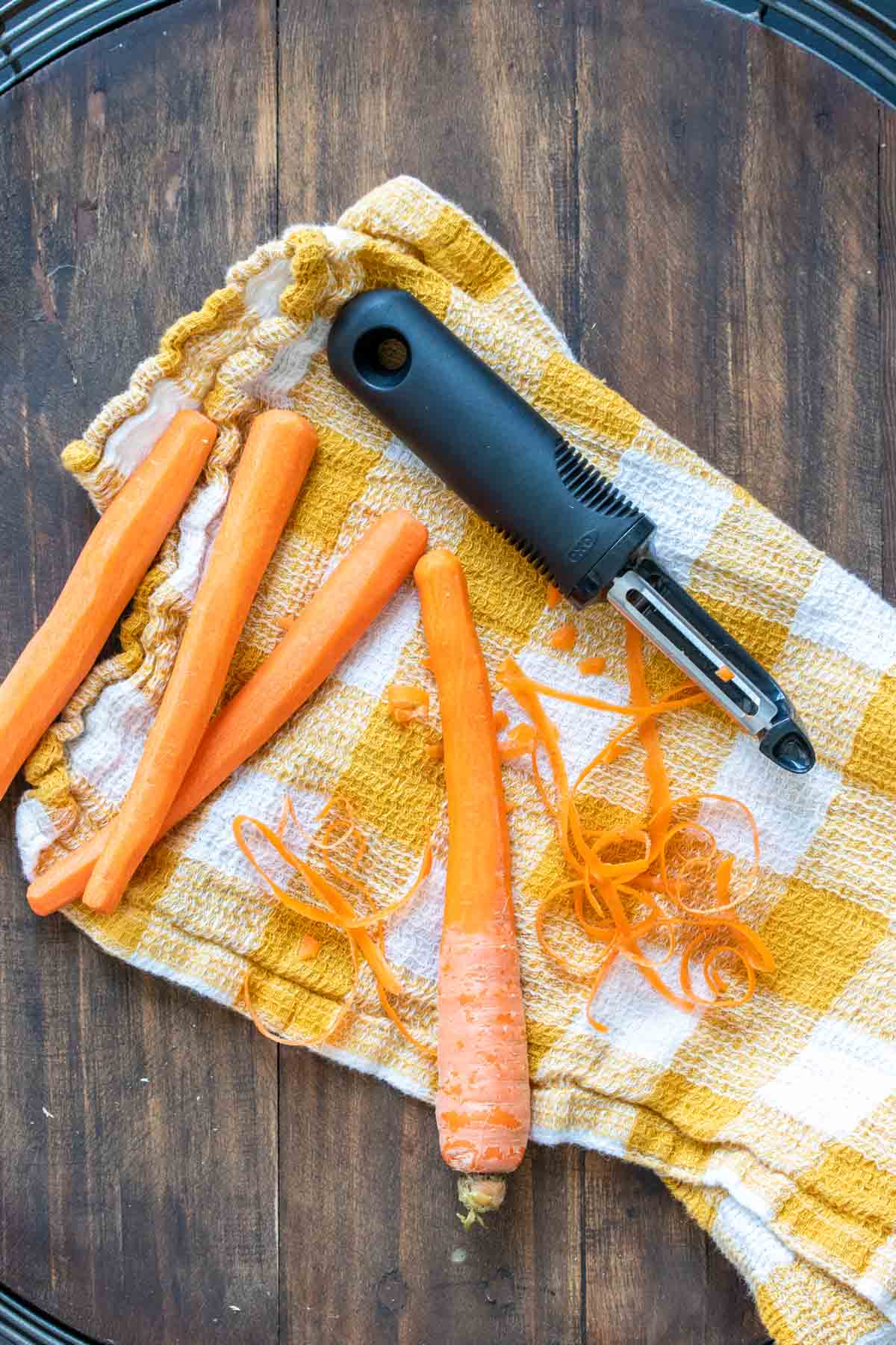 Carrots on a yellow checkered towel being peeled with a veggie peeler